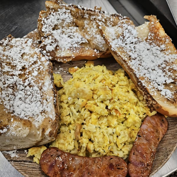 Cuban toast with powdered sugar, scrambled eggs, and two sausages on a wooden board.