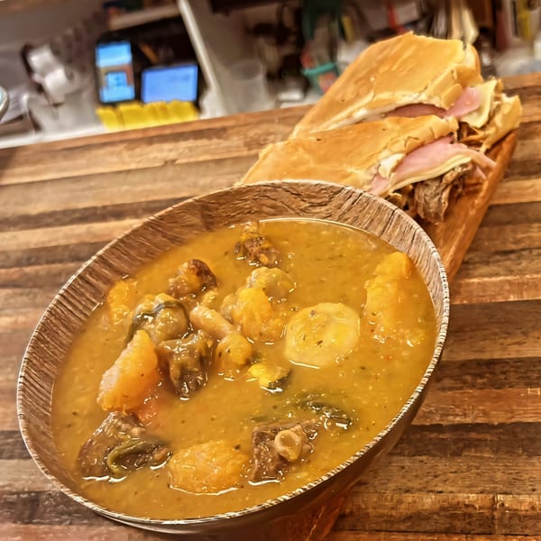 A bowl of Mima's Cuban Cafe's hearty stew with meat and vegetables, next to a Cuban sandwich.
